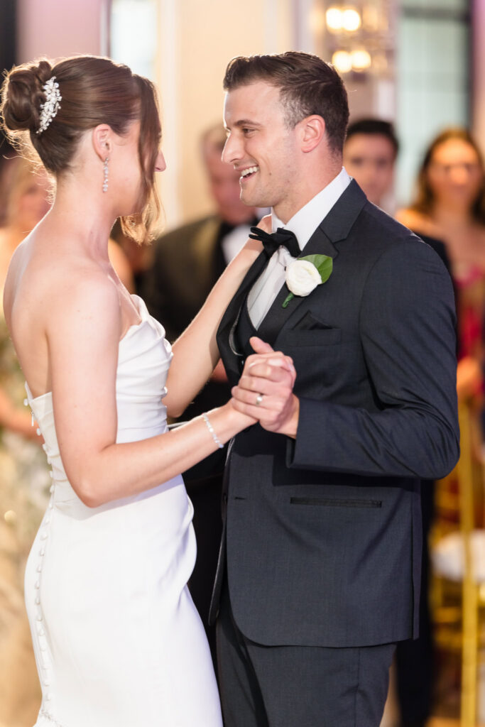 Tori and Mark hold hands in the Swan Club ballroom surrounded by floral centerpieces and floor-to-ceiling windows.