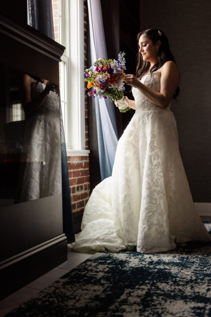 Bride in a floral wedding gown admiring her colorful bouquet beside a window before her Harbor Club at Prime wedding.
