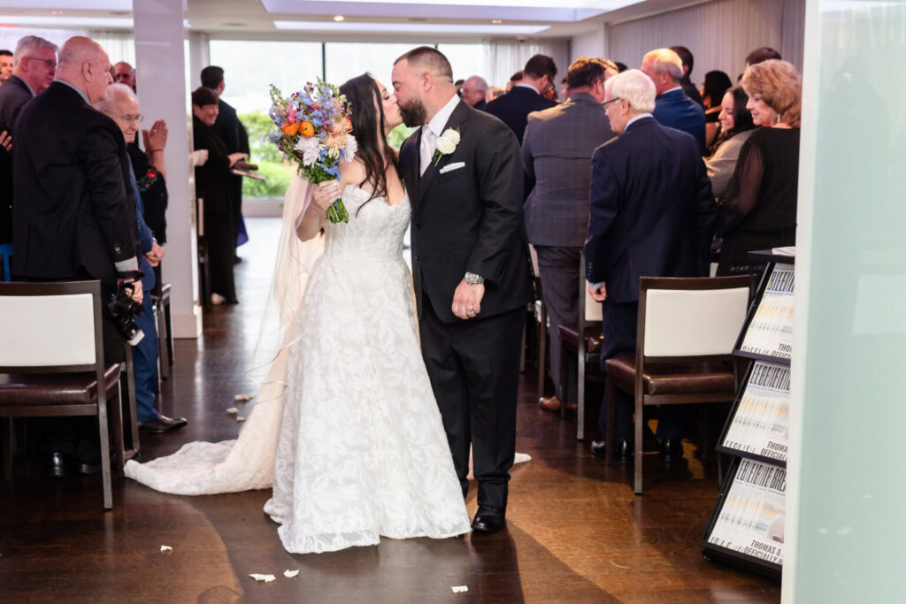 Newly married couple kissing as they walk down the aisle between wedding guests following their indoor ceremony.
