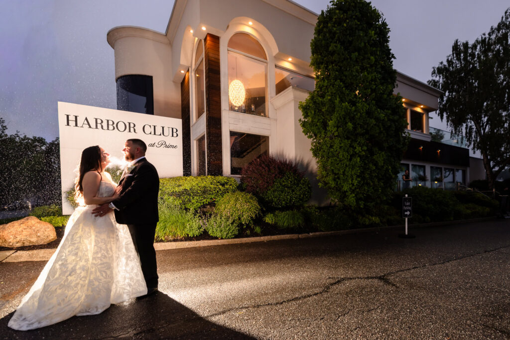 Bride and groom embracing outside Harbor Club at Prime at dusk, beside the venue sign and illuminated waterfront building.