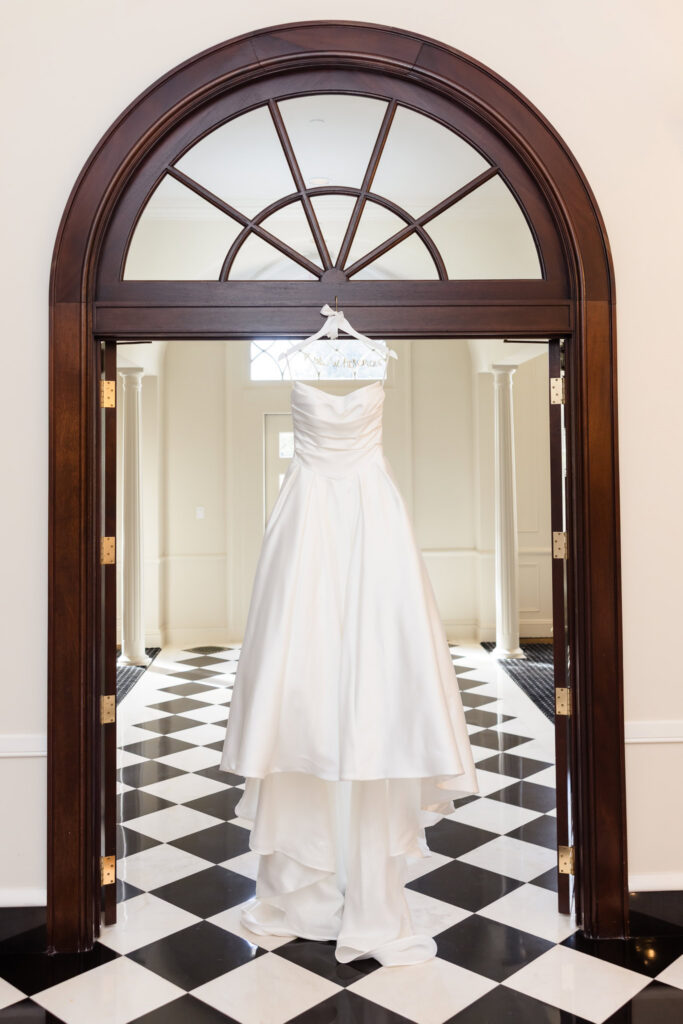 Strapless white wedding gown hanging beneath a dark wooden archway above black-and-white checkered flooring.