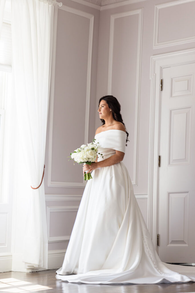 Morgan standing beside a softly lit window in her off-the-shoulder wedding gown, holding a white floral bouquet.