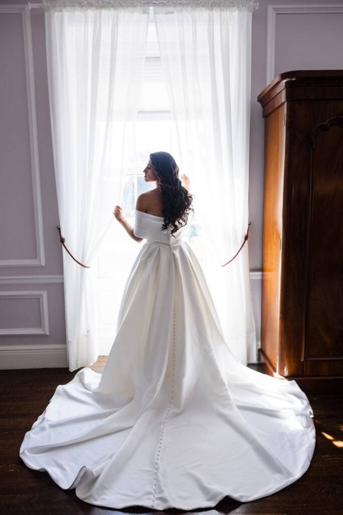 Rear portrait of Morgan standing by a window, showing the button detailing and sweeping train of her wedding gown.
