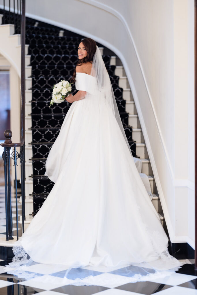 Morgan posing on a curved staircase in her wedding gown and veil, holding a bouquet of white flowers.