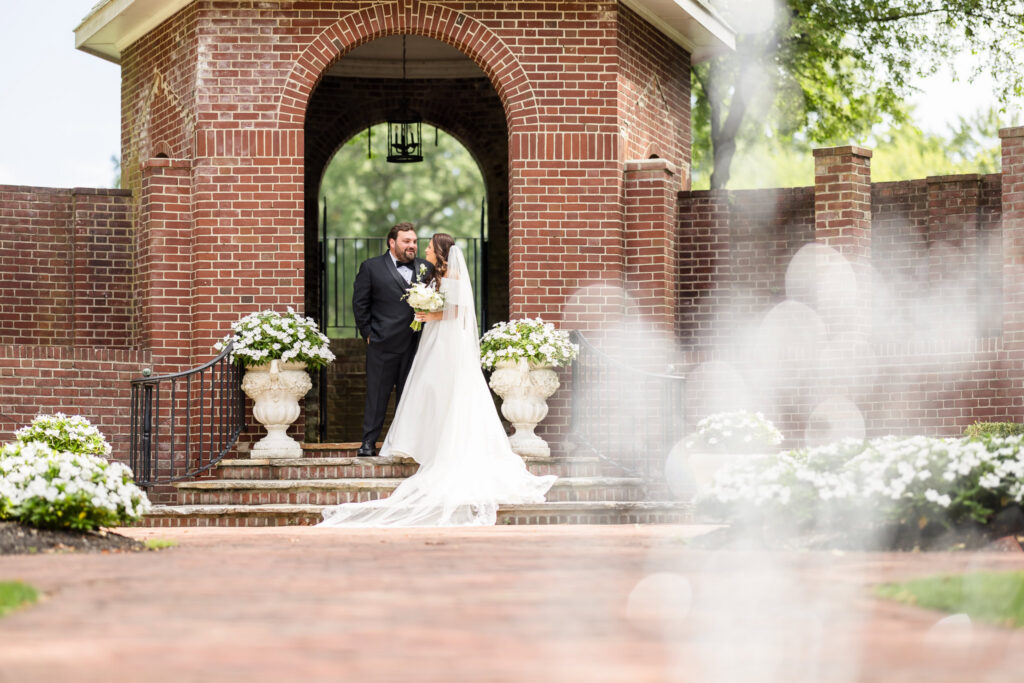 Bride and groom standing together beneath a brick garden archway surrounded by white flowers at The Mansion at Woodside wedding.