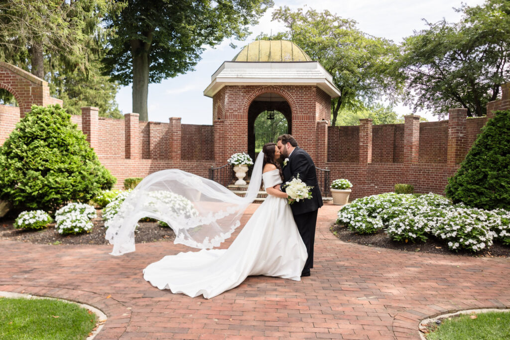 Bride and groom kissing in a brick courtyard as the bride's long veil flows behind them in the breeze.