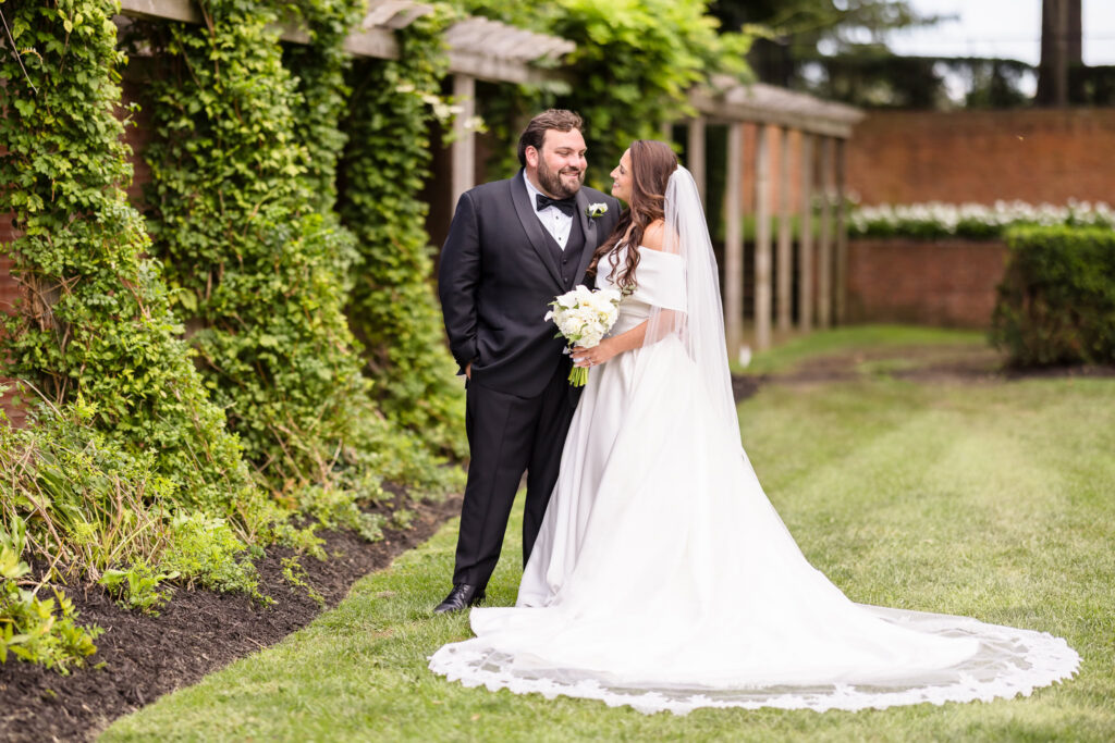 Bride in an off-the-shoulder wedding gown holding white flowers beside the groom in a black tuxedo along a lush garden walkway.