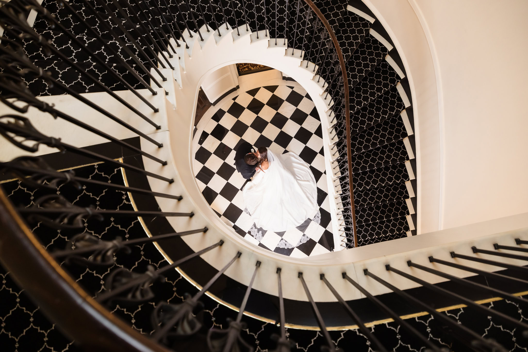 Overhead portrait of Morgan and James embracing beneath a curved staircase with black-and-white checkered flooring at The Mansion at Woodside.