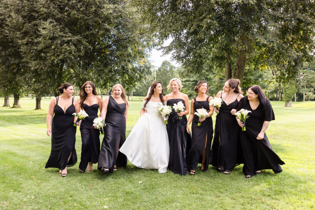 Bride laughing with seven bridesmaids dressed in elegant black gowns while walking together across a sunlit lawn.