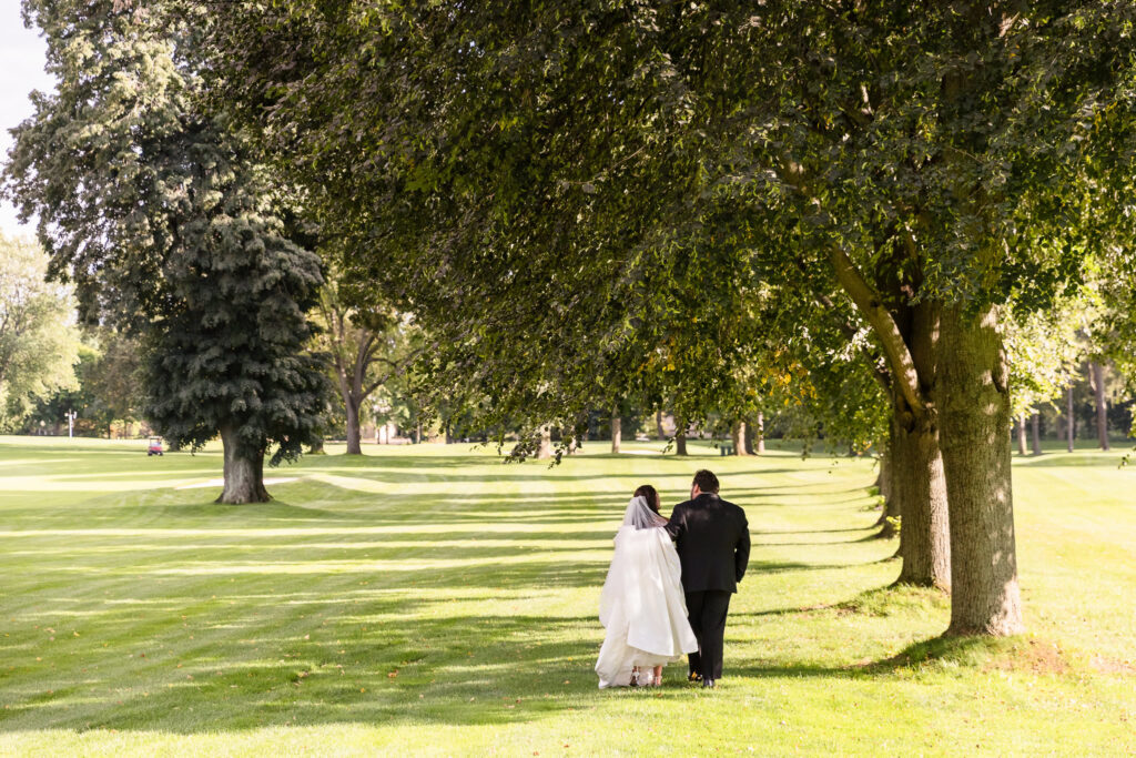 Bride and groom walking arm in arm beneath mature trees across a sunlit green lawn after their wedding ceremony.