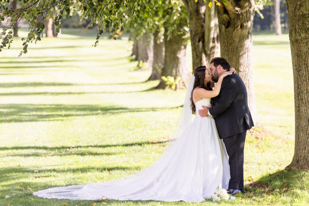Bride and groom sharing a romantic kiss beneath a tree, with the bride's long wedding gown and veil flowing across the grass.