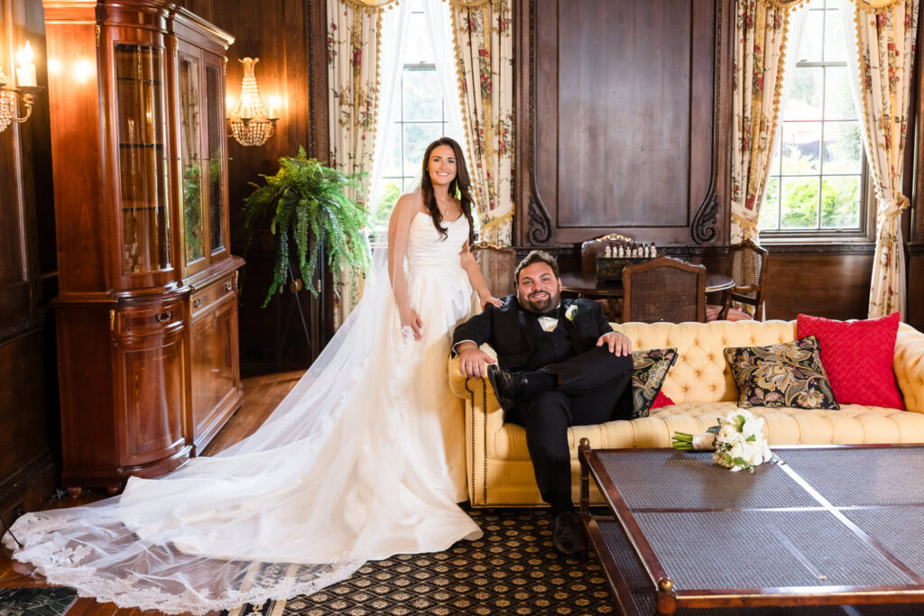 Bride standing beside the groom seated on a cream tufted sofa in an elegant wood-paneled room with floral curtains and antique furnishings.