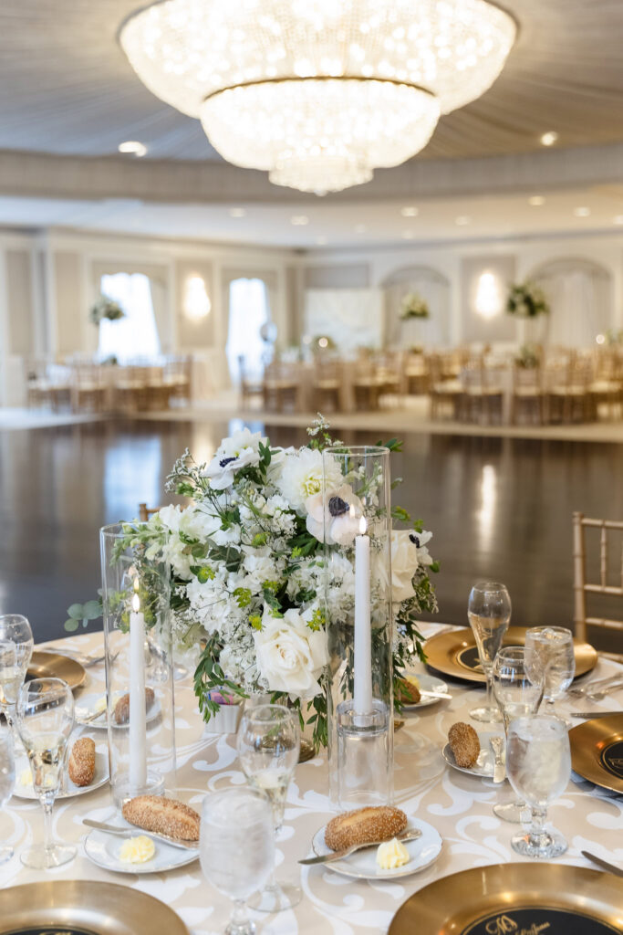 White floral wedding centerpiece with greenery and glass-enclosed taper candles on a reception table beneath a grand crystal chandelier.
