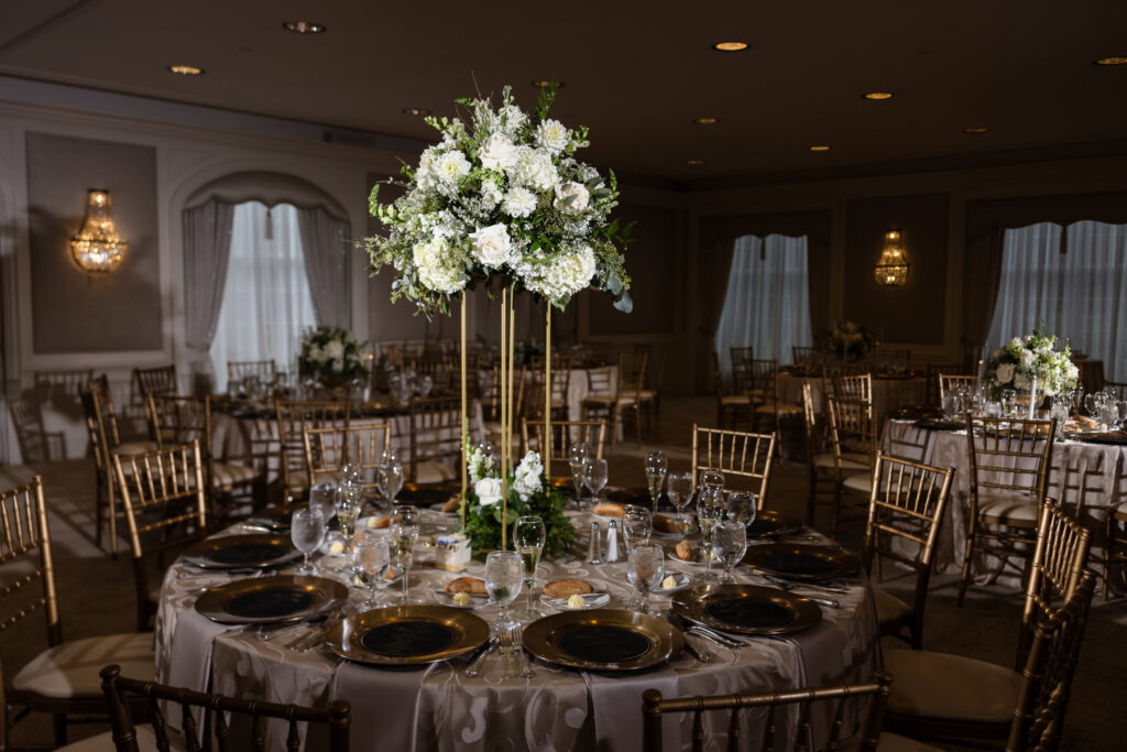 Tall white floral centerpiece displayed on a round reception table with gold chairs and place settings at The Mansion at Woodside.