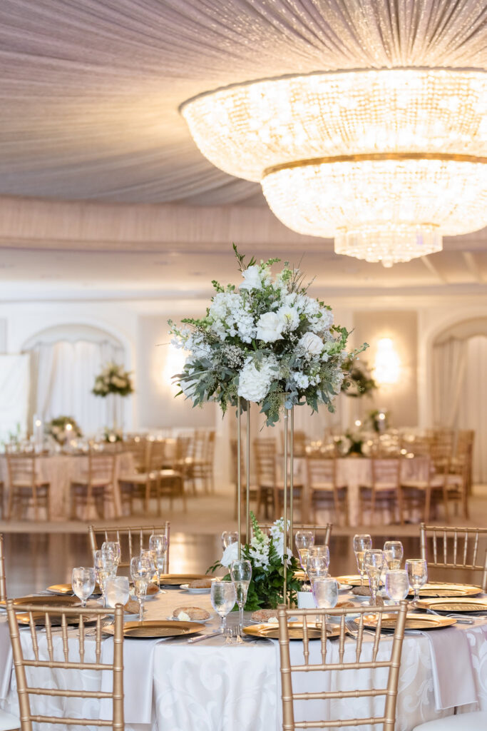 Elevated white floral centerpiece above a formal reception table with gold chairs and a sparkling crystal chandelier overhead.
