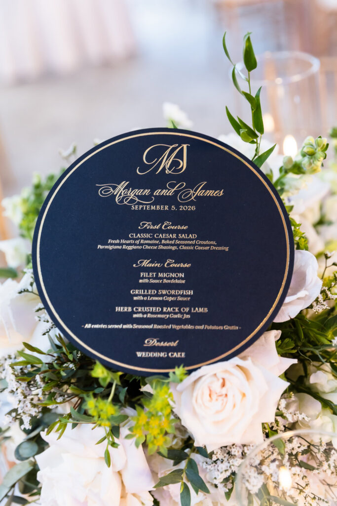 Round navy and gold wedding dinner menu for Morgan and James nestled among white roses and greenery at their reception.