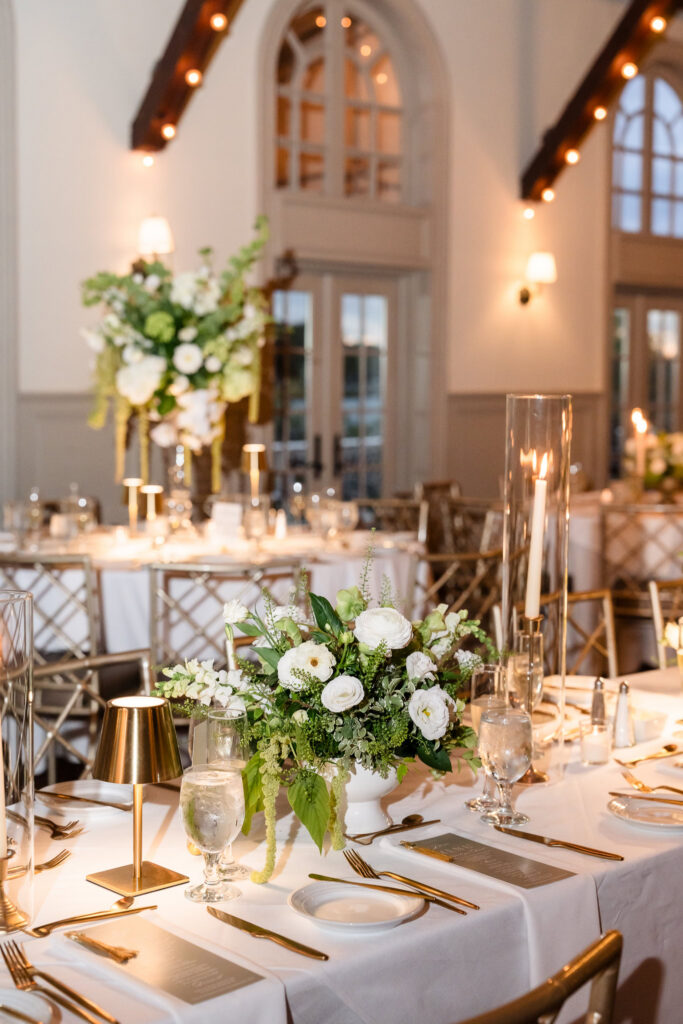 White and green floral centerpiece, candlelight, gold table lamps, and place settings inside the Old Field Club ballroom.