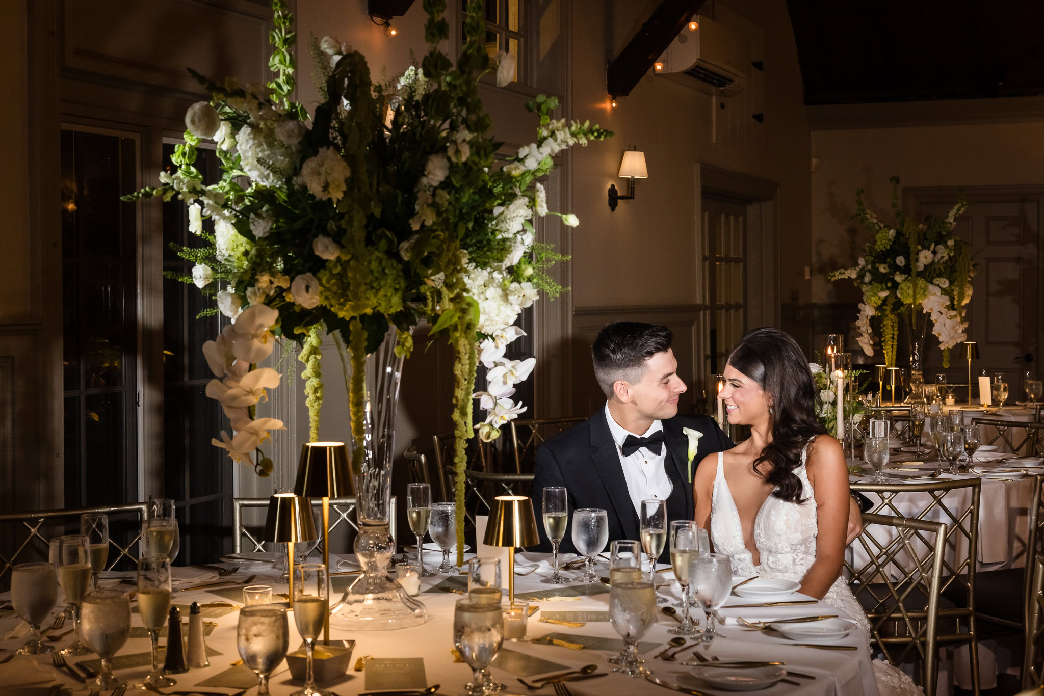 Bride and groom smiling at each other beside a tall floral centerpiece during their Old Field Club wedding reception.