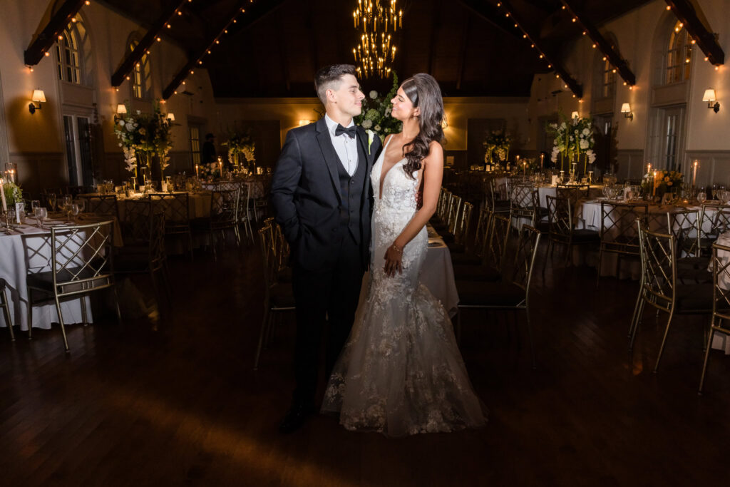 Bride and groom facing each other beneath a chandelier in the warmly lit Old Field Club ballroom.