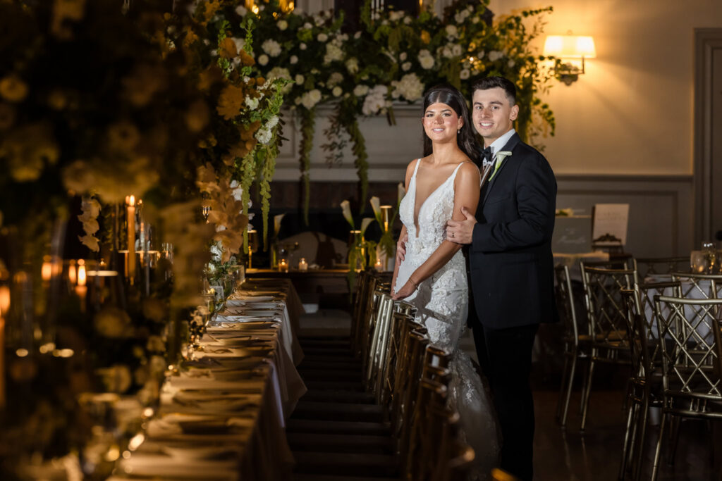 Bride and groom posing beside candlelit reception tables and white floral arrangements at the Old Field Club.