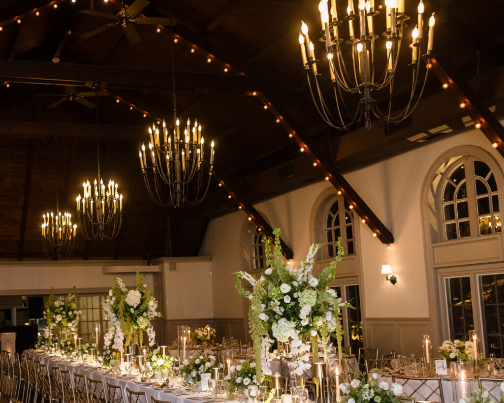 Old Field Club ballroom with vaulted wood ceiling, chandeliers, arched windows, and reception tables decorated with white flowers.