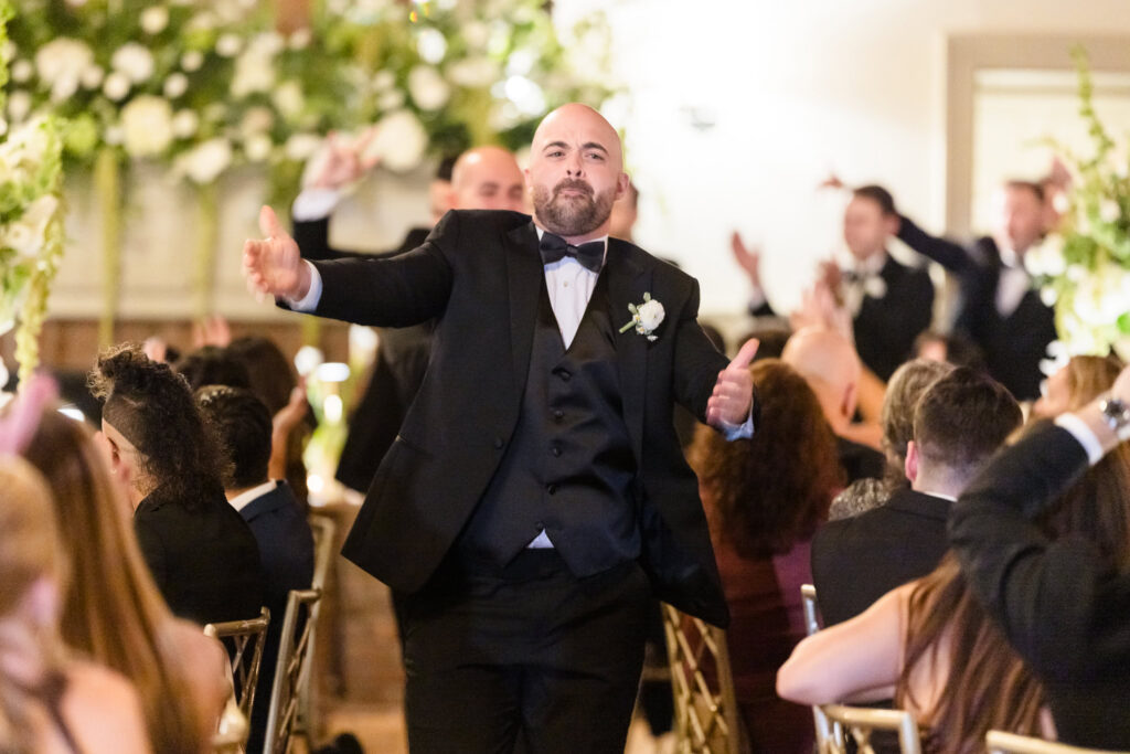 Wedding guest in a black tuxedo celebrating with raised arms among seated guests at the Old Field Club reception.